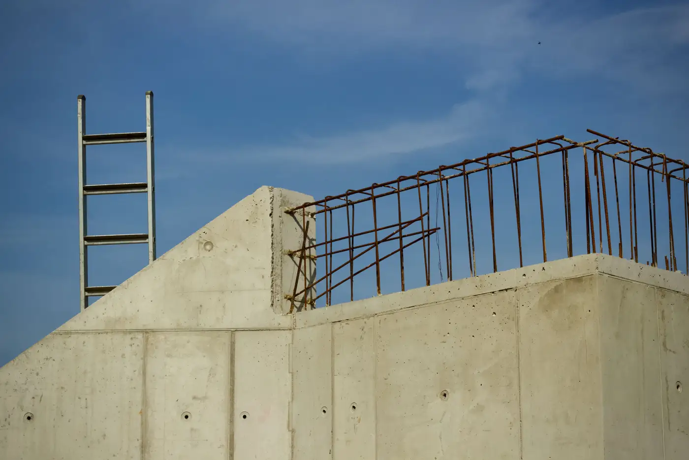 Top of a poured concrete wall with a ladder and exposed rebar stubs against a blue sky.