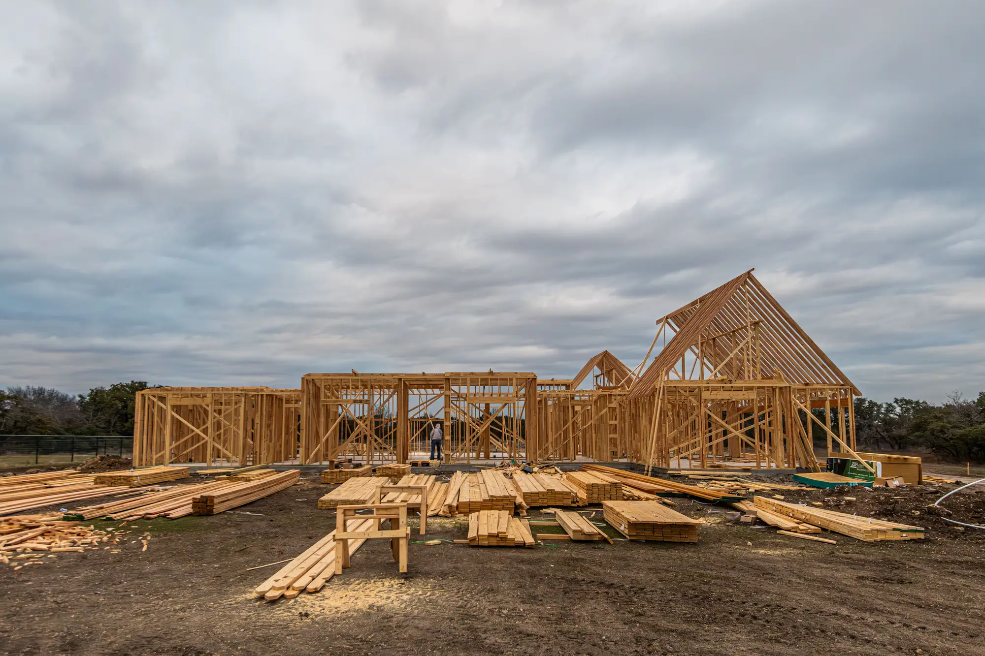 Timber framing for a house going up under an overcast sky, lumber stacked in front