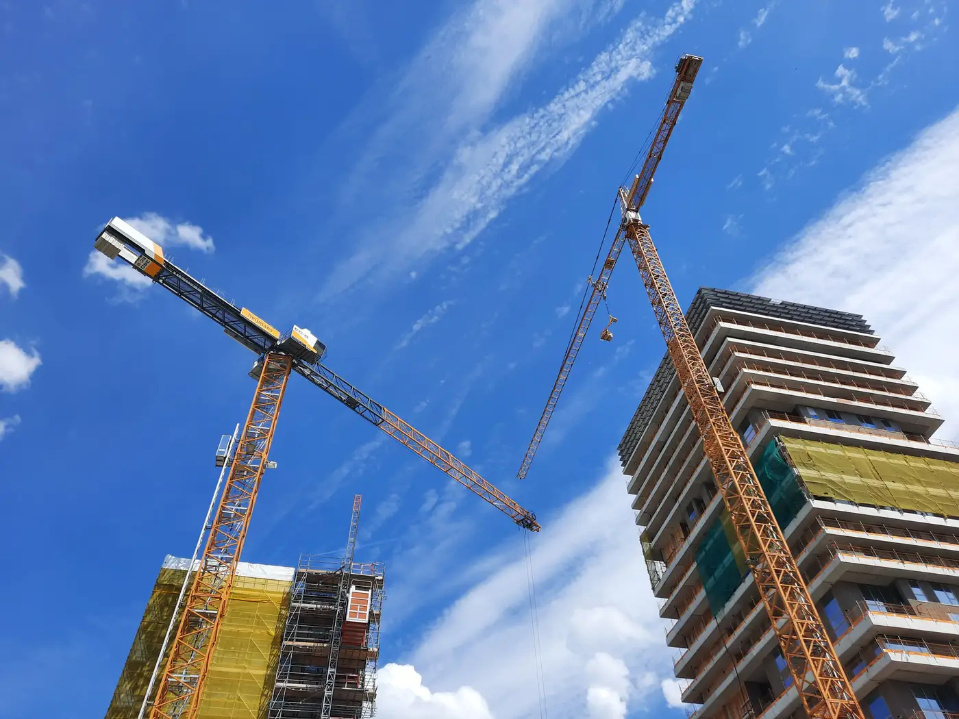 Two tower cranes over a mid-rise concrete building under construction against a blue sky.